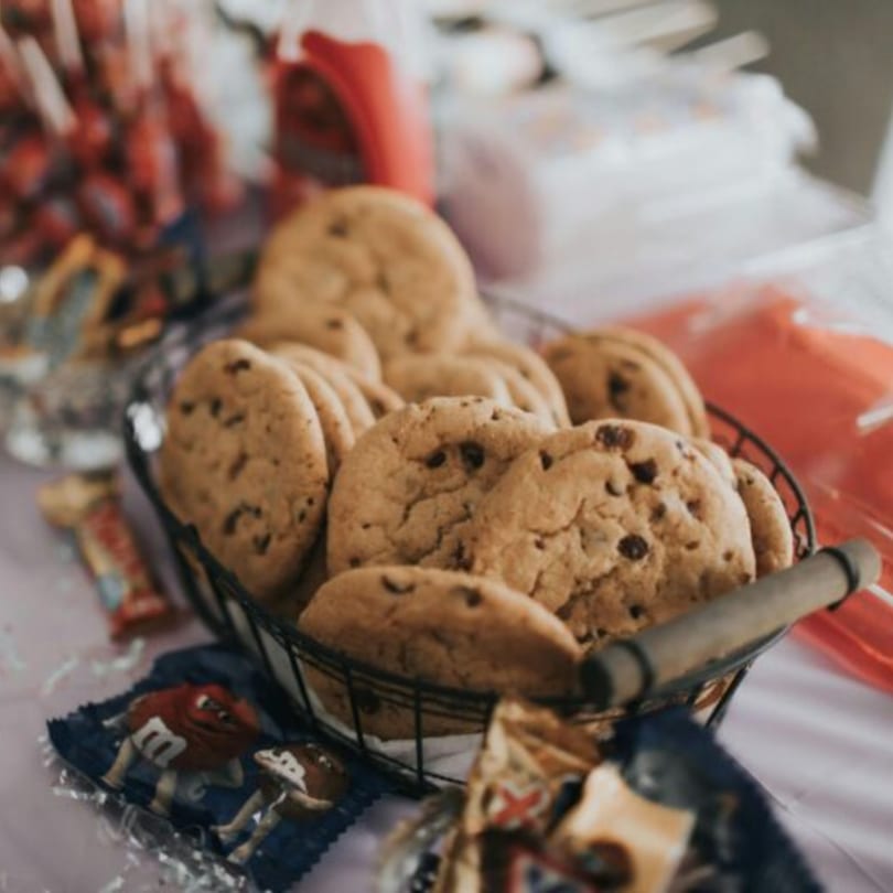 A wire basket filled with chocolate chip cookies, with candy wrappers scattered around.