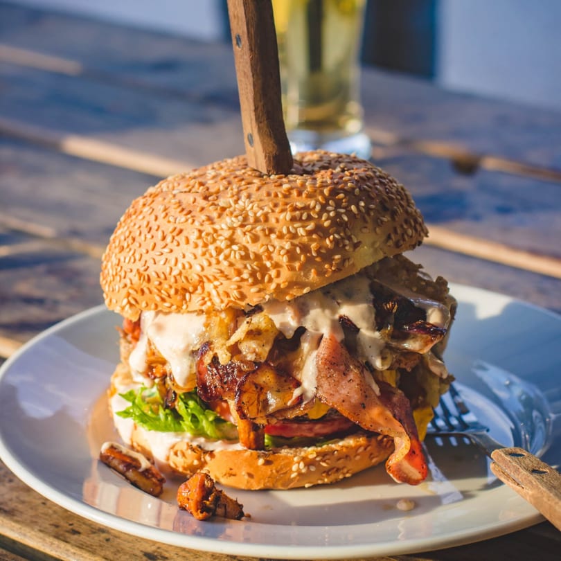 A large burger with bacon, sauce, and vegetables, held together by a knife, on a white plate.